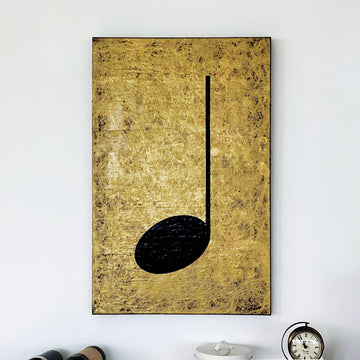 Gold and black music themed painting hanging on a white wall above some wine bottles and a clock